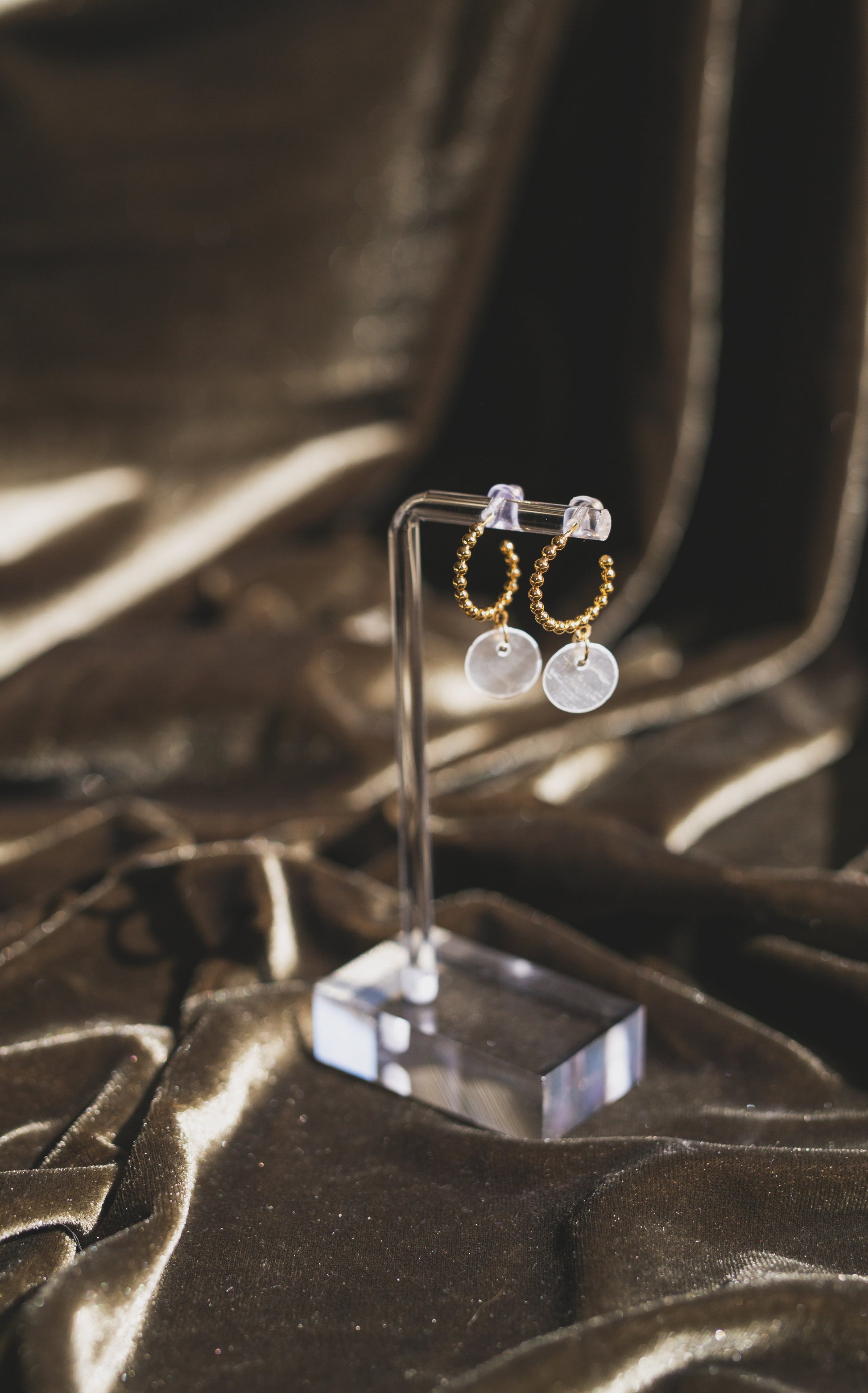 A pair of gold hoop earrings with round white disc charms and small clear stone studs, displayed on a clear acrylic stand against a draped dark brown satin fabric background.